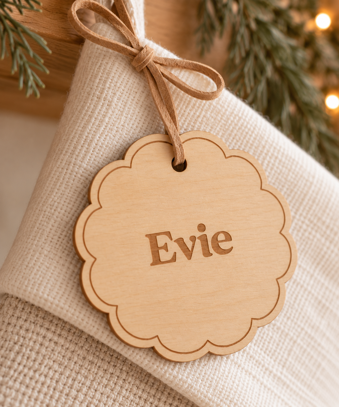 A scalloped wooden ornament tag engraved with the name 'Evie', hanging from a twine bow on a textured cream fabric, with blurred evergreen branches and warm string lights in the background.