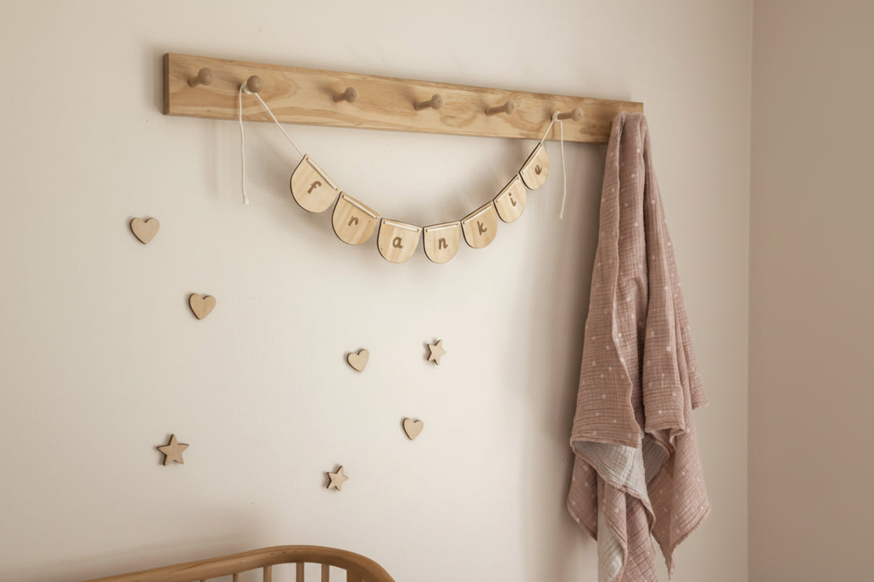 Wooden crib with a wooden name bunting hung from wall hooks and pink towel on a white wall.