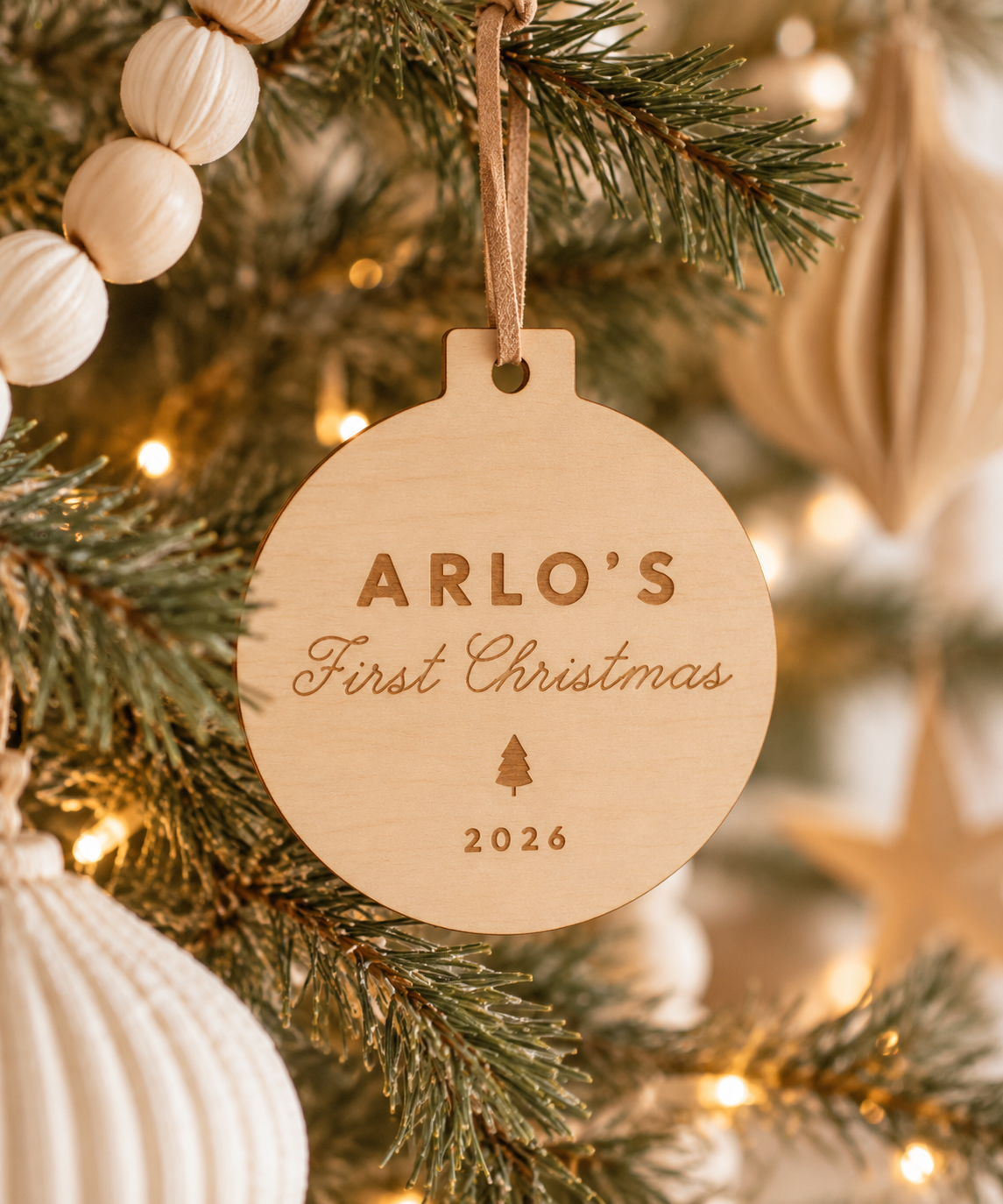 A personalized round wooden Christmas ornament hanging from a pine tree branch with warm string lights. The ornament is engraved with the text 'ARLO'S First Christmas' and the year '2026' in a natural light wood finish.