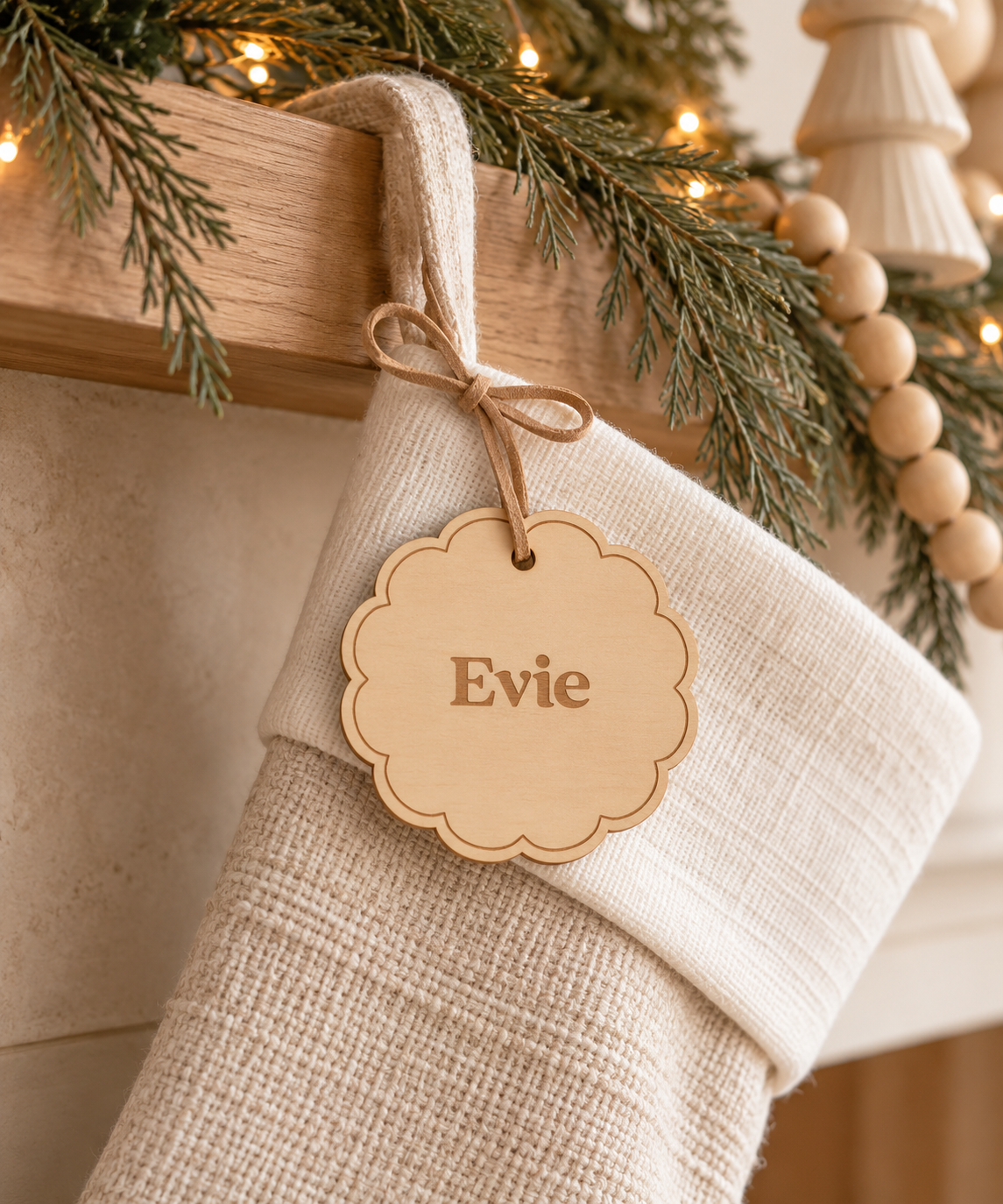 A beige linen Christmas stocking hanging from a wooden mantel, decorated with a scalloped natural wood tag engraved with the name 'Evie'. The scene is styled with a green pine garland, warm string lights, and wooden beads.